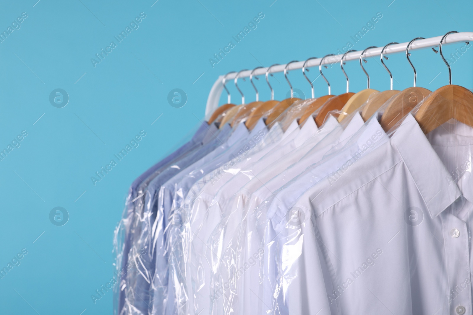 Photo of Dry-cleaning service. Many different clothes in plastic bags hanging on rack against light blue background, closeup and space for text