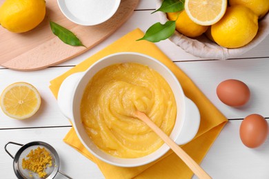 Delicious lemon curd in bowl, ingredients, spoon and sieve on white wooden table, flat lay