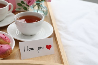 Photo of Tasty breakfast served in bed. Delicious eclairs, tea and card with phrase I Love You on tray, closeup. Space for text