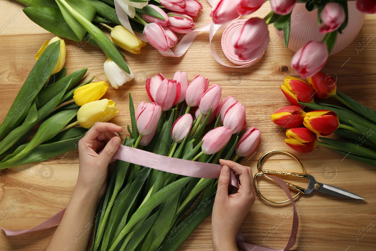 Photo of Woman making beautiful bouquet of fresh tulips and ribbon at wooden table, top view