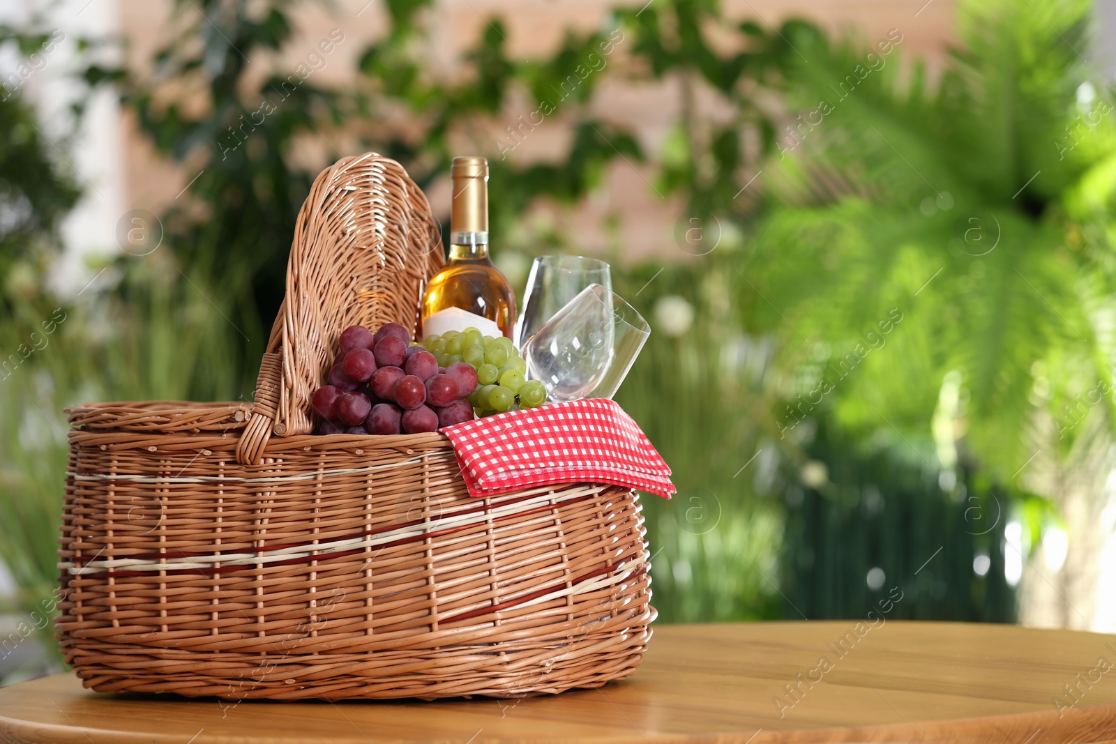 Photo of Picnic basket with wine, glasses and grapes on wooden table against blurred background, space for text