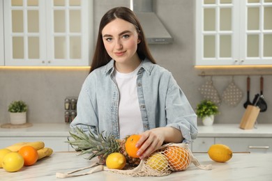 Photo of Woman with string bag of fresh fruits at light marble table in kitchen