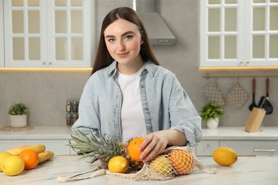 Woman with string bag of fresh fruits at light marble table in kitchen