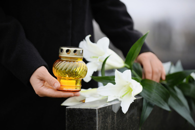 Photo of Woman with candle and white lilies near black granite tombstone outdoors, closeup. Funeral ceremony