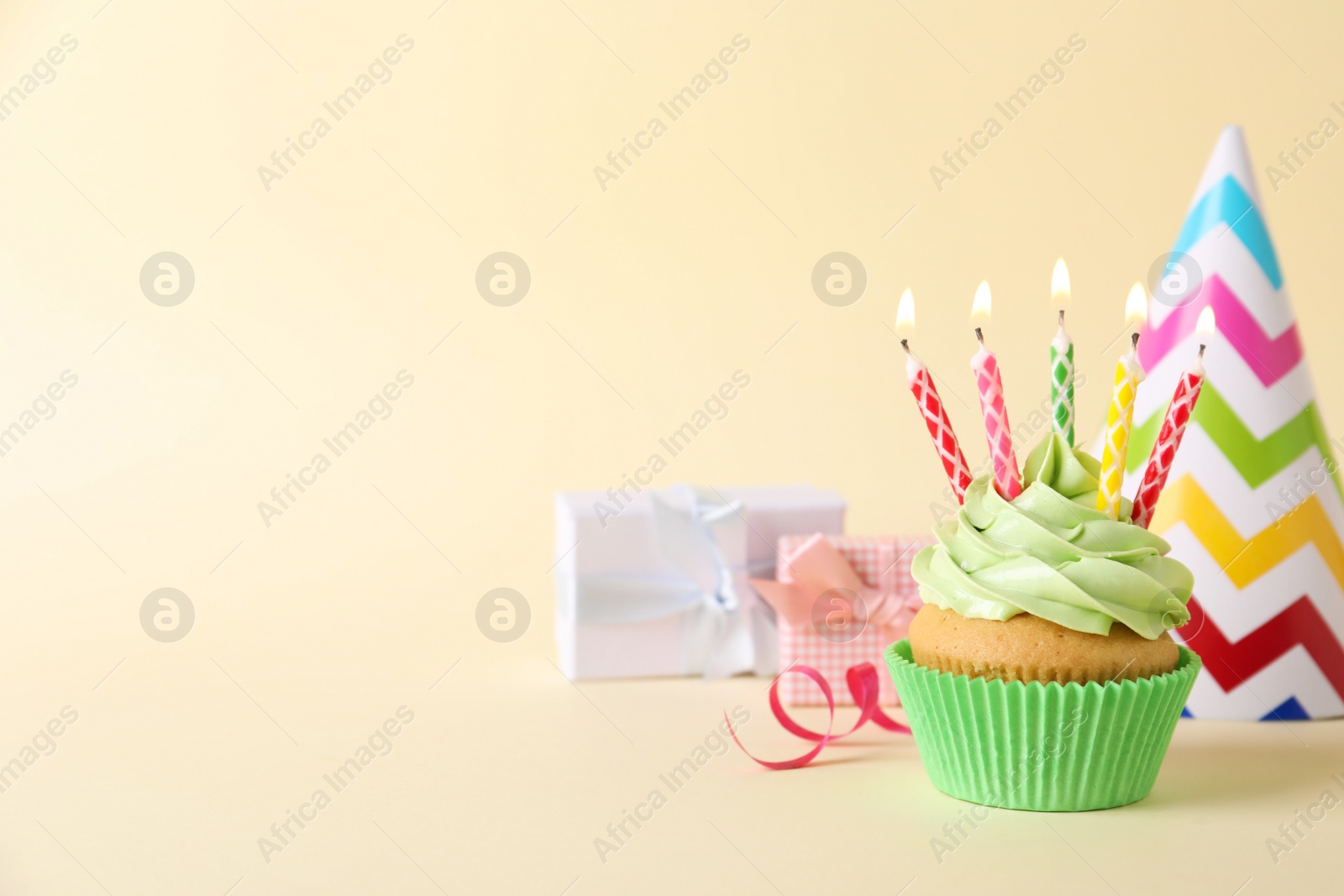 Photo of Birthday cupcake with burning candles, gift boxes and party hat on beige background. Space for text