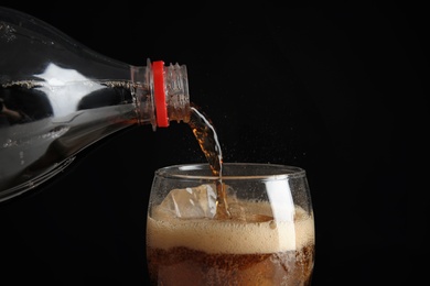 Pouring refreshing cola from bottle into glass with ice cubes on black background, closeup