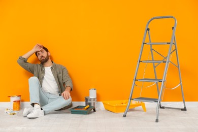 Photo of Tired designer sitting on floor near freshly painted orange wall indoors