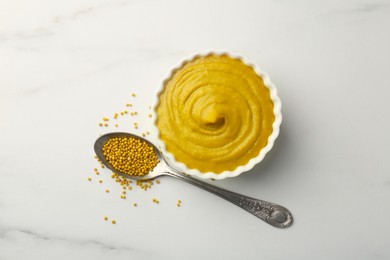 Photo of Tasty mustard sauce in bowl and spoon with dry seeds on white marble table, flat lay