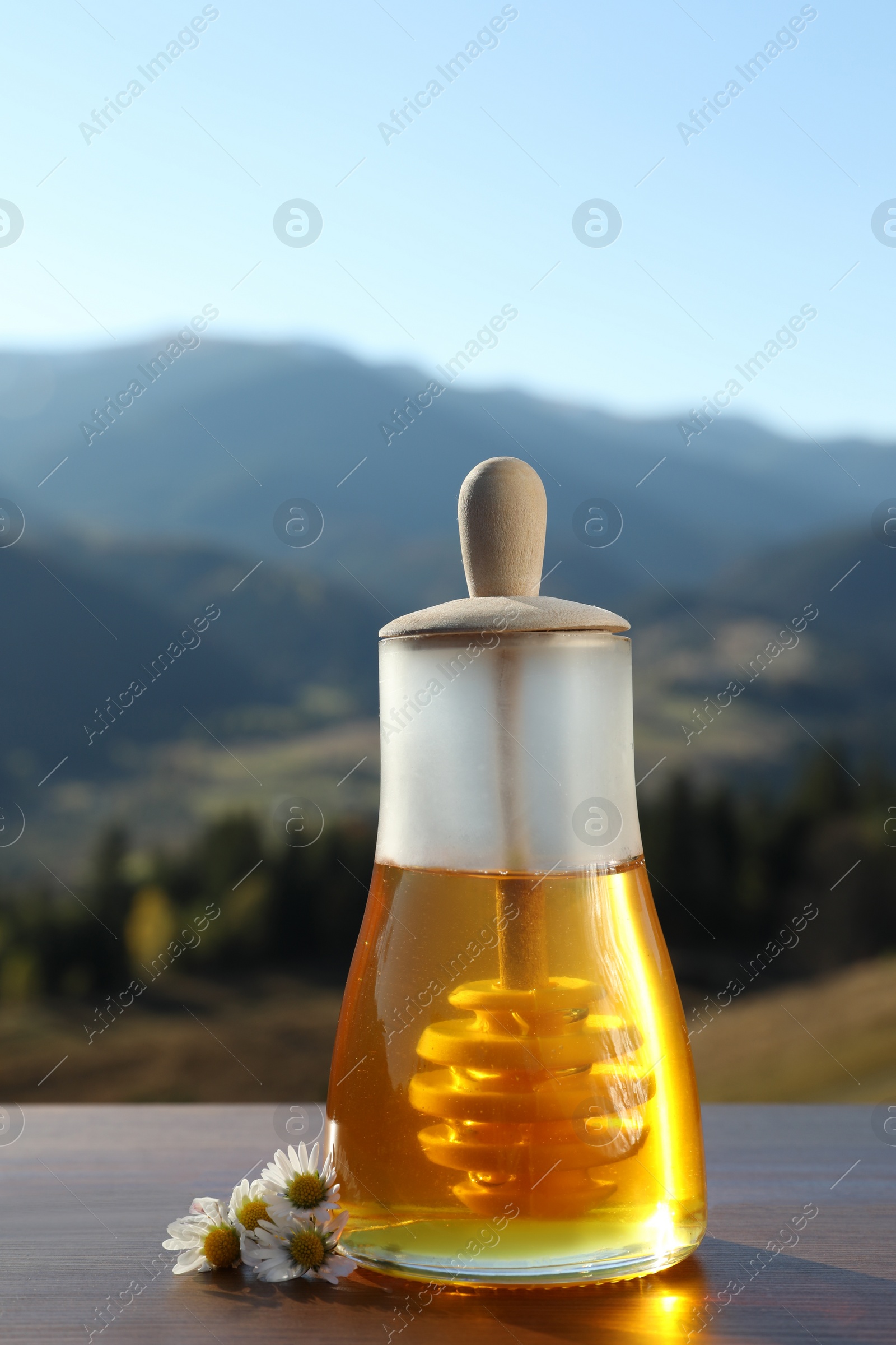 Photo of Fresh aromatic honey and chamomile flowers on wooden table against mountain landscape