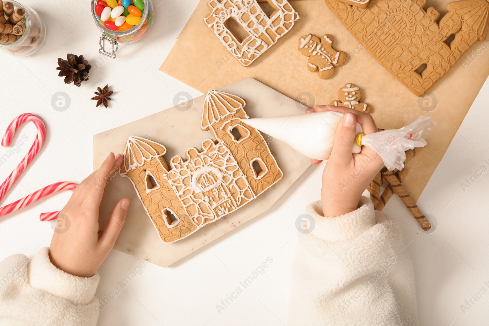 Photo of Woman decorating gingerbread house part with icing at white table, top view