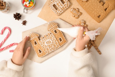 Woman decorating gingerbread house part with icing at white table, top view