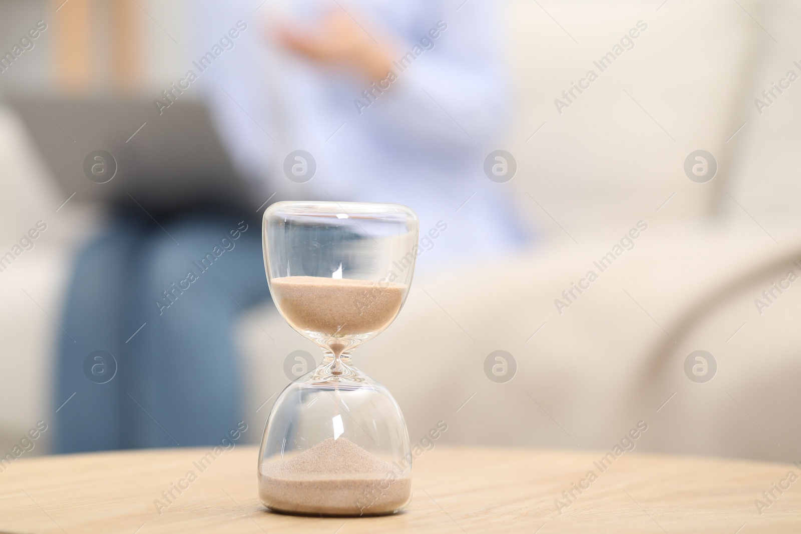 Photo of Hourglass with flowing sand on desk. Woman using laptop indoors, selective focus