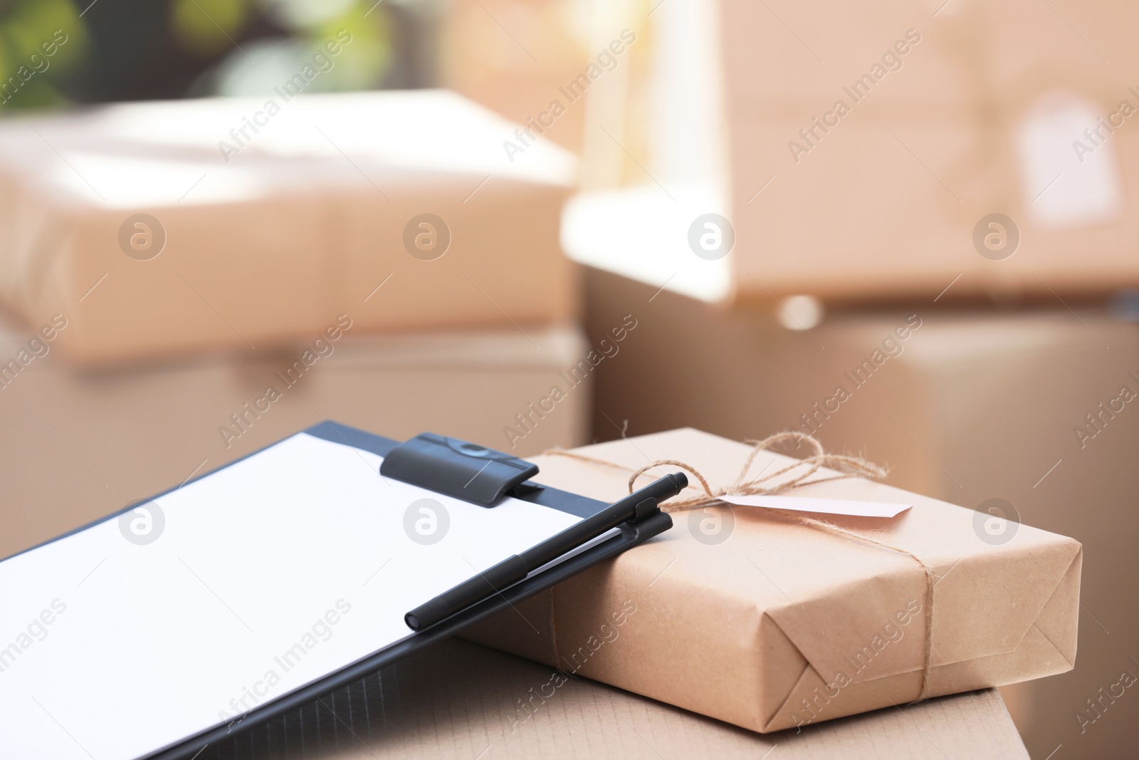 Photo of Clipboard, parcel with tag and blurred stacked boxes on background, indoors