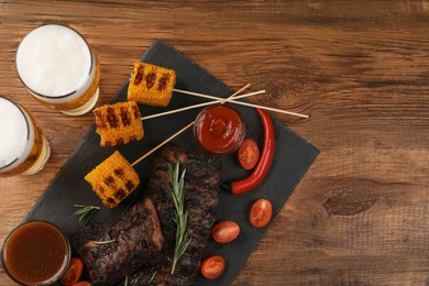 Glasses of beer, delicious grilled ribs, corn and sauces on wooden table, flat lay. Space for text