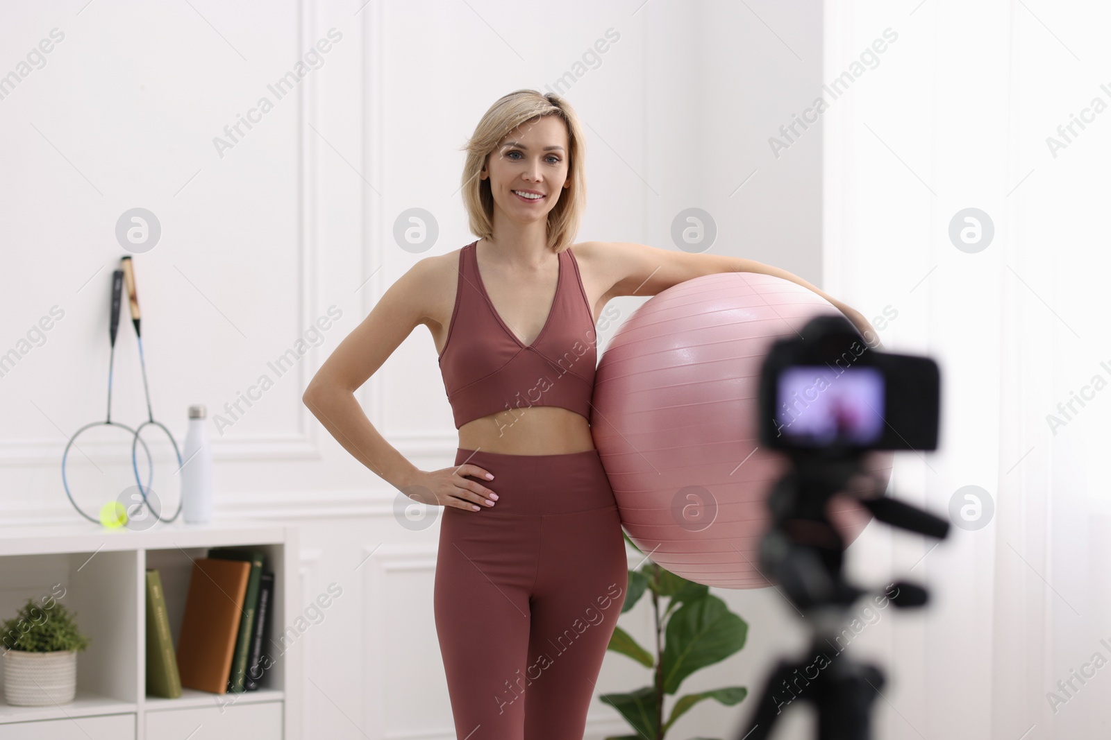 Photo of Smiling sports blogger holding fit ball while recording fitness lesson with camera at home