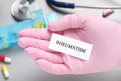 Photo of Doctor in glove holding sheet of paper with word Rheumatism above grey table, closeup