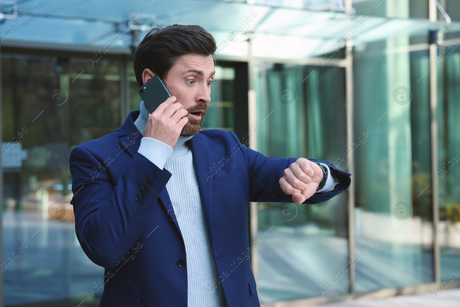 Photo of Emotional businessman talking on smartphone and looking at watch near building. Being late concept