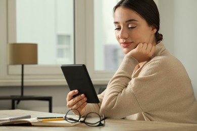 Photo of Young woman using e-book reader at wooden table indoors