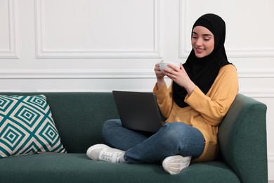 Muslim woman in hijab with cup of drink using laptop on sofa indoors. Space for text