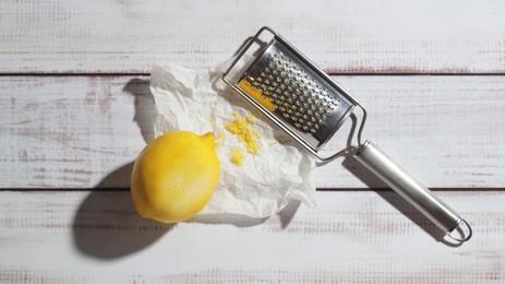 Photo of Lemon, grater and paper with fresh zest on white wooden table, top view