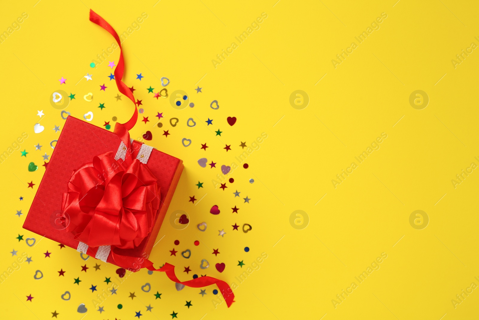 Photo of Red gift box and shiny confetti on yellow background, top view. Space for text