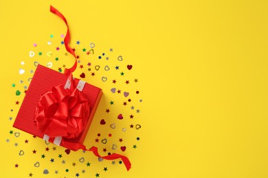 Red gift box and shiny confetti on yellow background, top view. Space for text