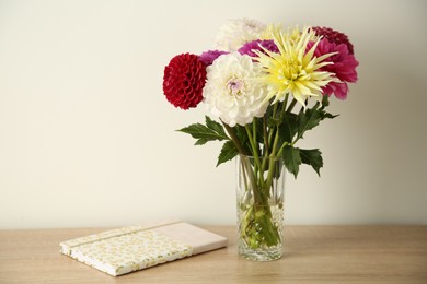 Photo of Bouquet of beautiful Dahlia flowers in vase and notebook on wooden table near white wall