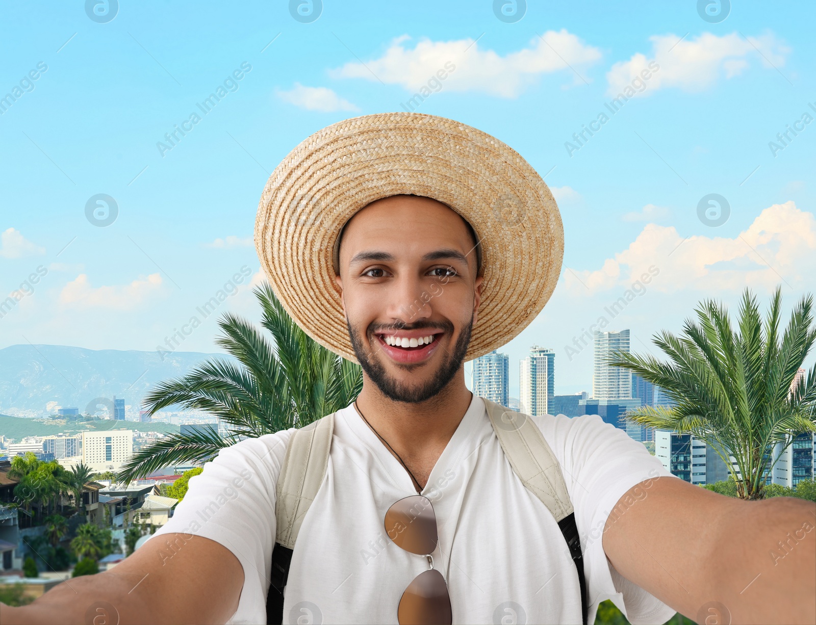 Image of Smiling young man in straw hat taking selfie against city with buildings and beautiful palms
