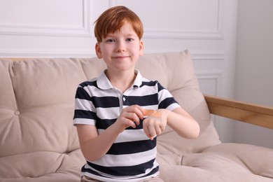 Little boy putting sticking plaster onto hand on sofa indoors