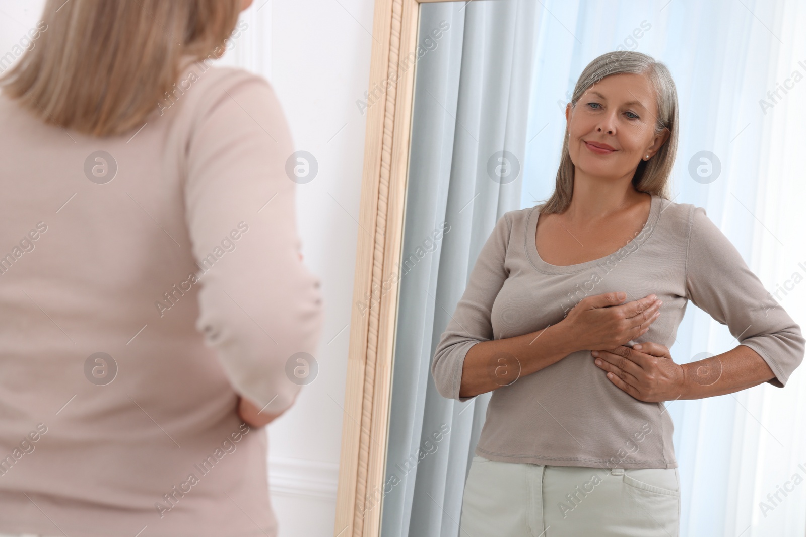 Photo of Beautiful senior woman doing breast self-examination near mirror indoors