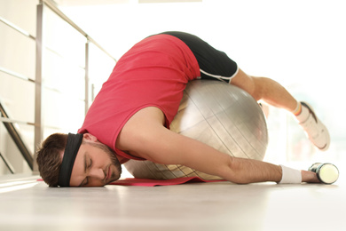 Lazy young man with exercise ball on yoga mat indoors