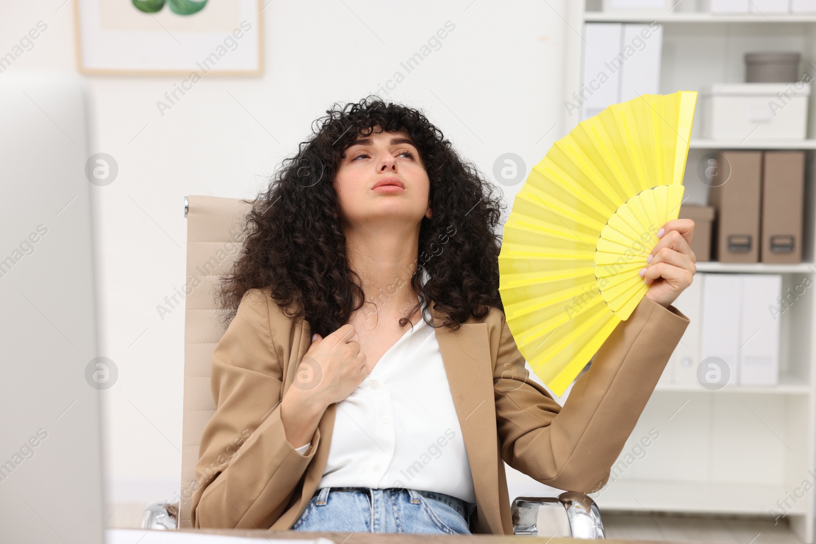 Photo of Young businesswoman waving yellow hand fan to cool herself in office