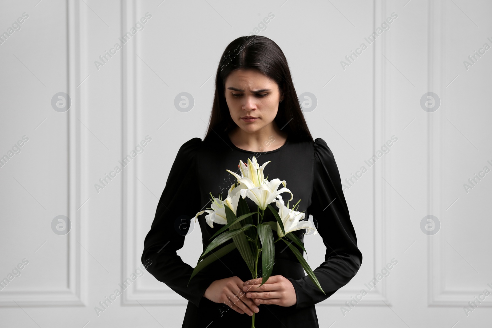 Photo of Sad woman with lilies mourning near white wall. Funeral ceremony