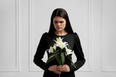 Photo of Sad woman with lilies mourning near white wall. Funeral ceremony