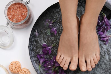 Photo of Woman soaking her feet in bowl with water and flowers on white wooden floor, top view. Spa treatment