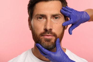 Photo of Doctor checking patient's face before cosmetic surgery operation on pink background, closeup