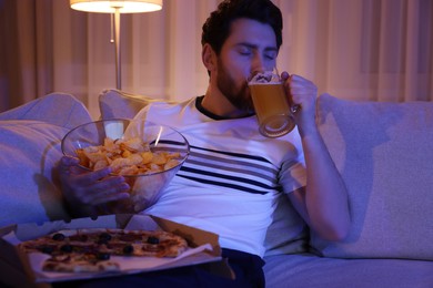 Photo of Man with chips, pizza and glass of beer on sofa at night. Bad habit