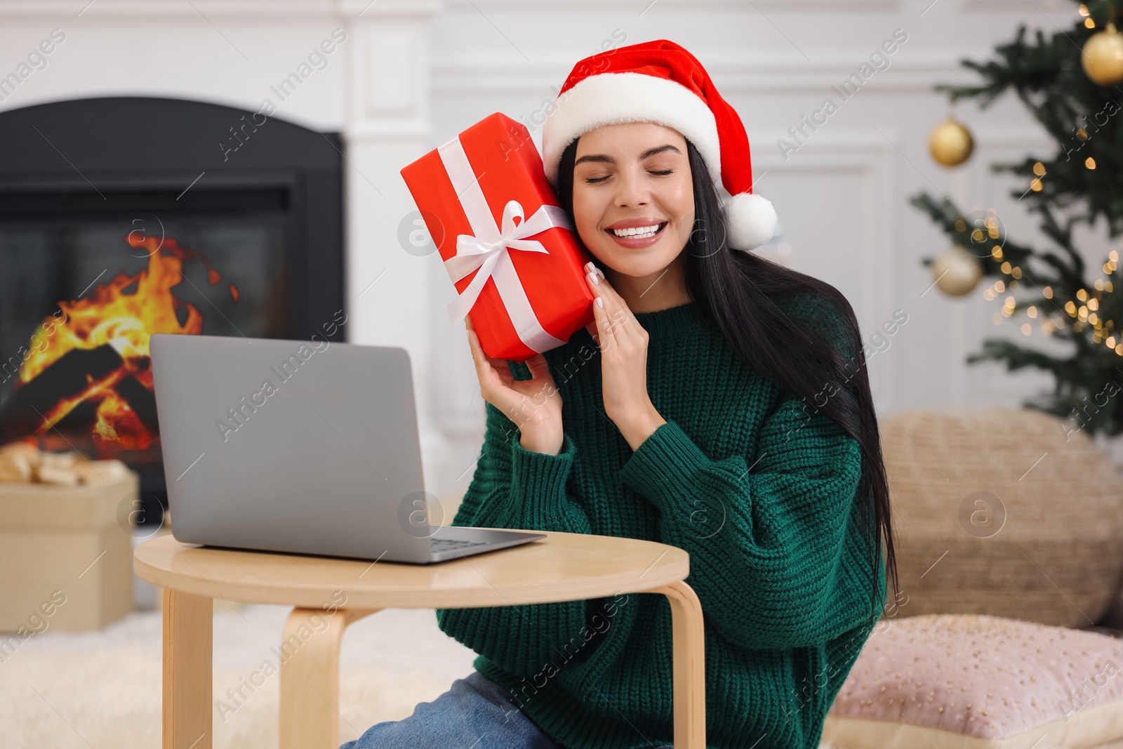 Photo of Celebrating Christmas online with exchanged by mail presents. Smiling woman in Santa hat with gift box during video call on laptop at home