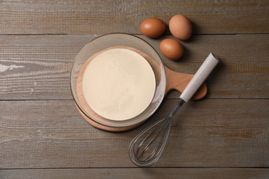 Photo of Metal whisk, dough in bowl and eggs on wooden table, flat lay