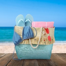 Image of Stylish bag with beach accessories on wooden surface near seashore 