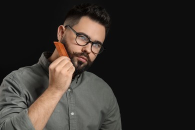 Photo of Handsome young man combing beard on black background. Space for text