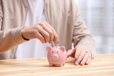 Photo of Man putting coin into piggy bank at wooden table, closeup