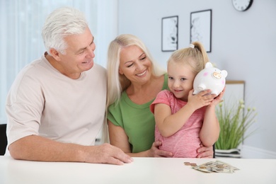 Photo of Little girl with piggy bank and her grandparents at table