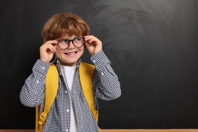 Photo of Happy schoolboy in glasses with backpack near blackboard, space for text