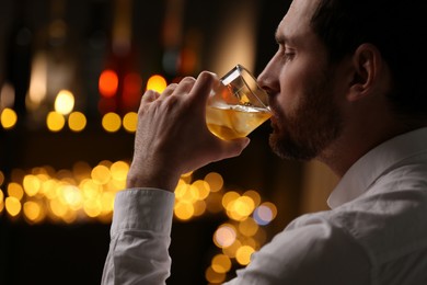 Handsome man drinking whiskey against blurred lights, closeup