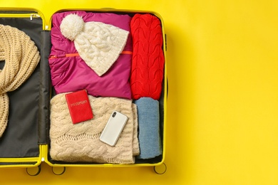 Open suitcase with warm clothes, passport and smartphone on color background, top view. Space for text