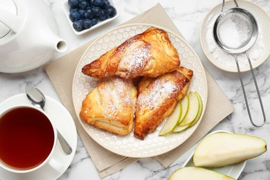 Photo of Fresh tasty puff pastry with sugar powder, pear, blueberries and aromatic tea served on white marble table, flat lay