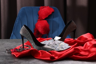 Prostitution concept. High heeled shoes, dollar banknotes, condom and handcuffs on grey textured table near chair with bra. Space for text