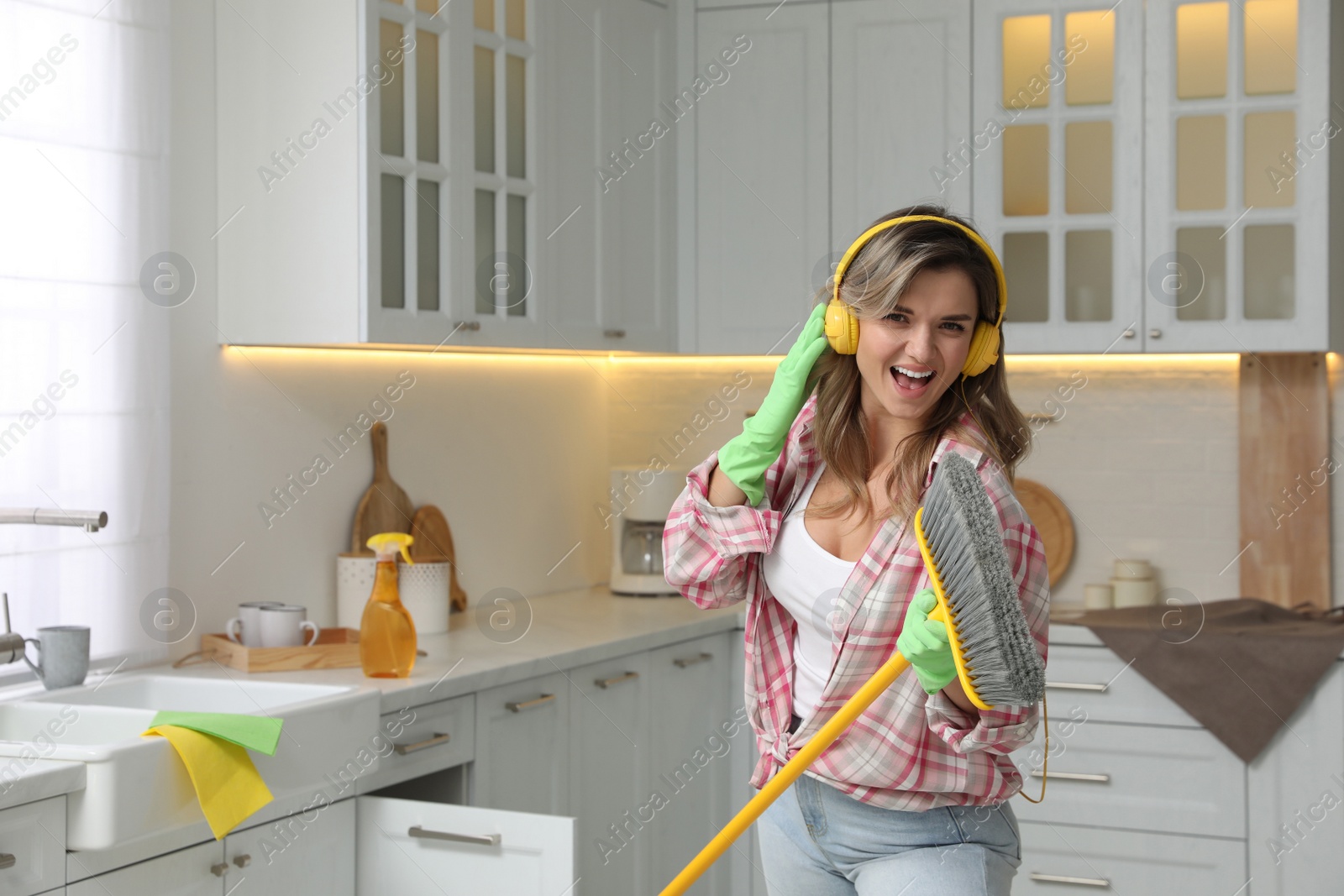 Photo of Beautiful young woman with headphones singing while cleaning kitchen