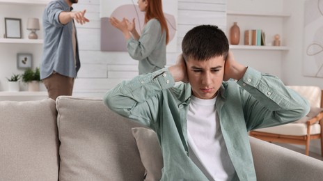 Photo of Unhappy teenage boy covering ears while his parents arguing on background. Problems at home